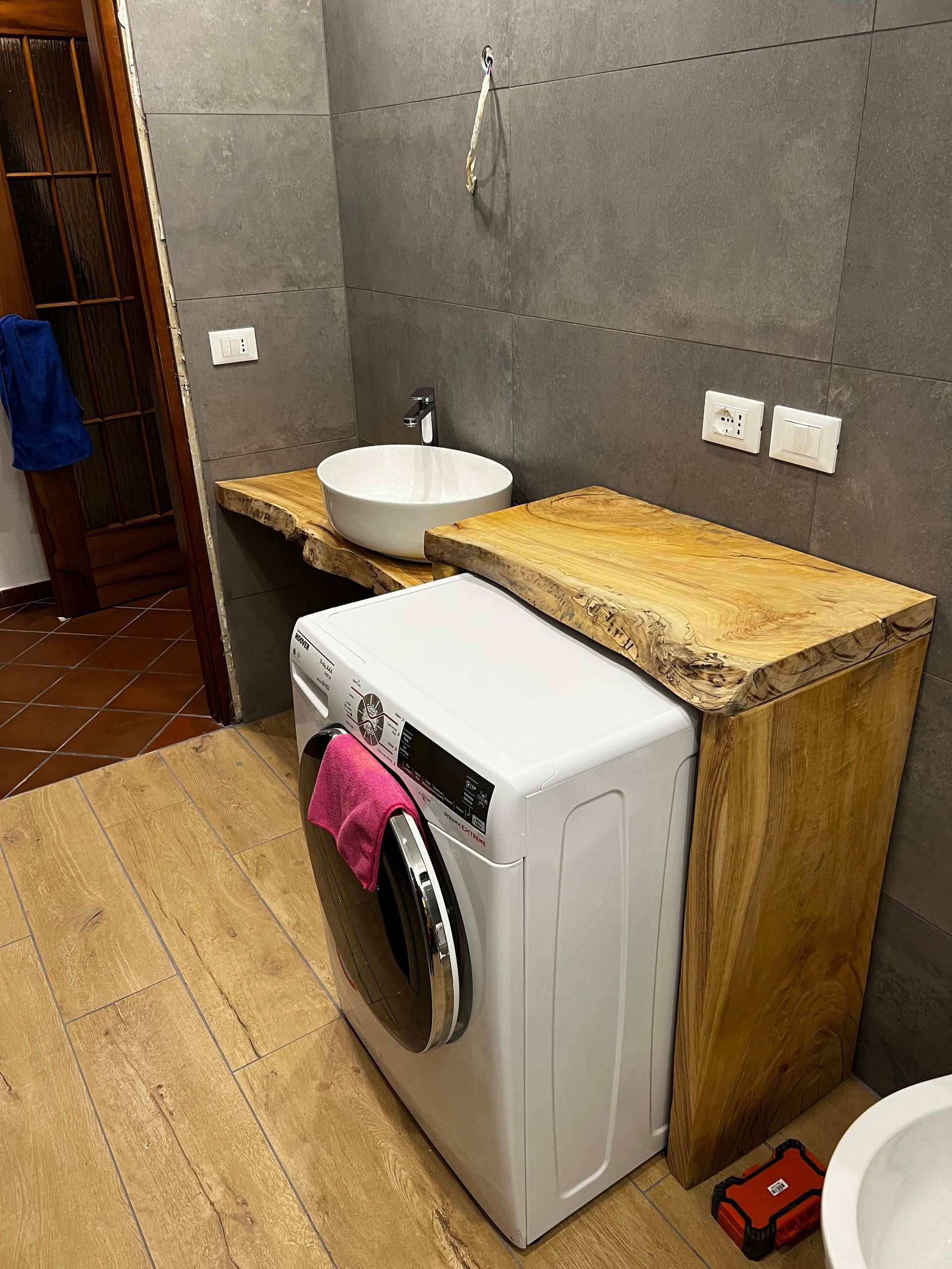 Bagno con lavatrice, lavandino e piano di lavoro in legno. Colori grigio e marrone chiaro.