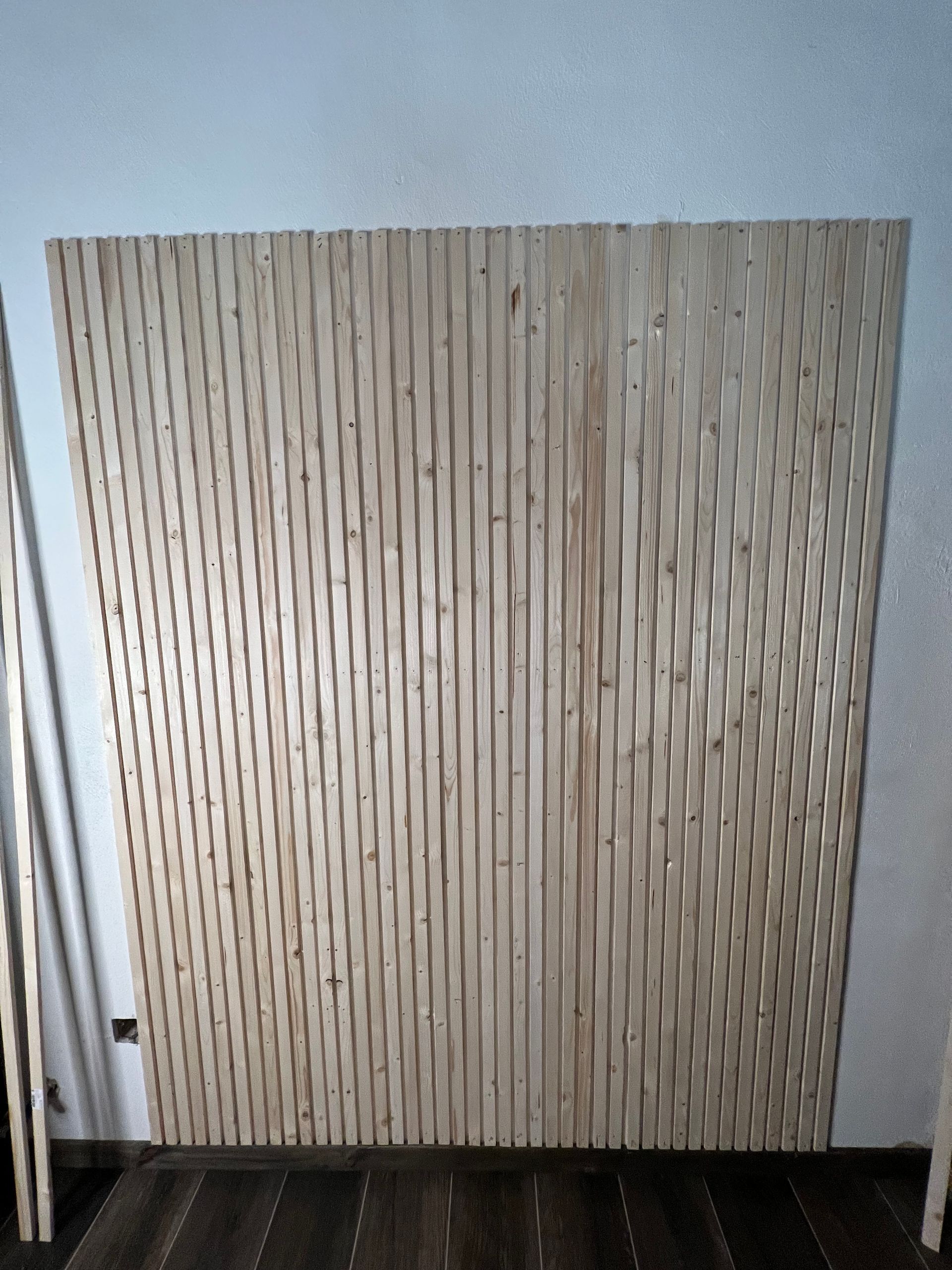 Parete con pannelli in legno con scanalature verticali, appoggiata a una parete bianca in una stanza con pavimento in legno.