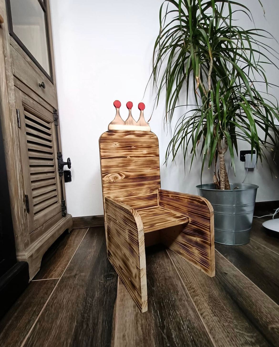 Sedia per bambini in legno con motivo a corona, all'interno di una stanza, vicino a un mobiletto e a una pianta in vaso.
