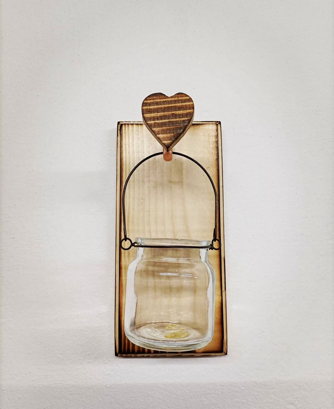 Porta-barattoli da parete in legno con dettaglio a forma di cuore. Un barattolo trasparente è appeso a un anello metallico su una tavola di legno tinto.