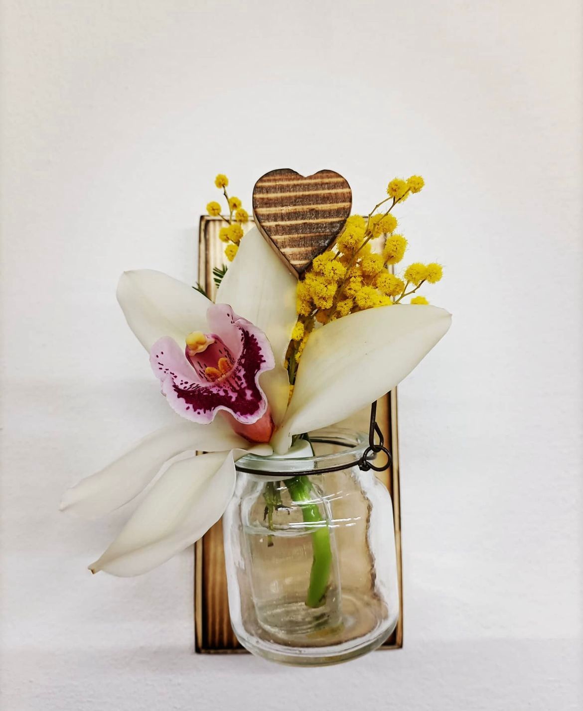 Composizione floreale con orchidea, mimosa e cuore di legno in un piccolo barattolo, su sfondo bianco.