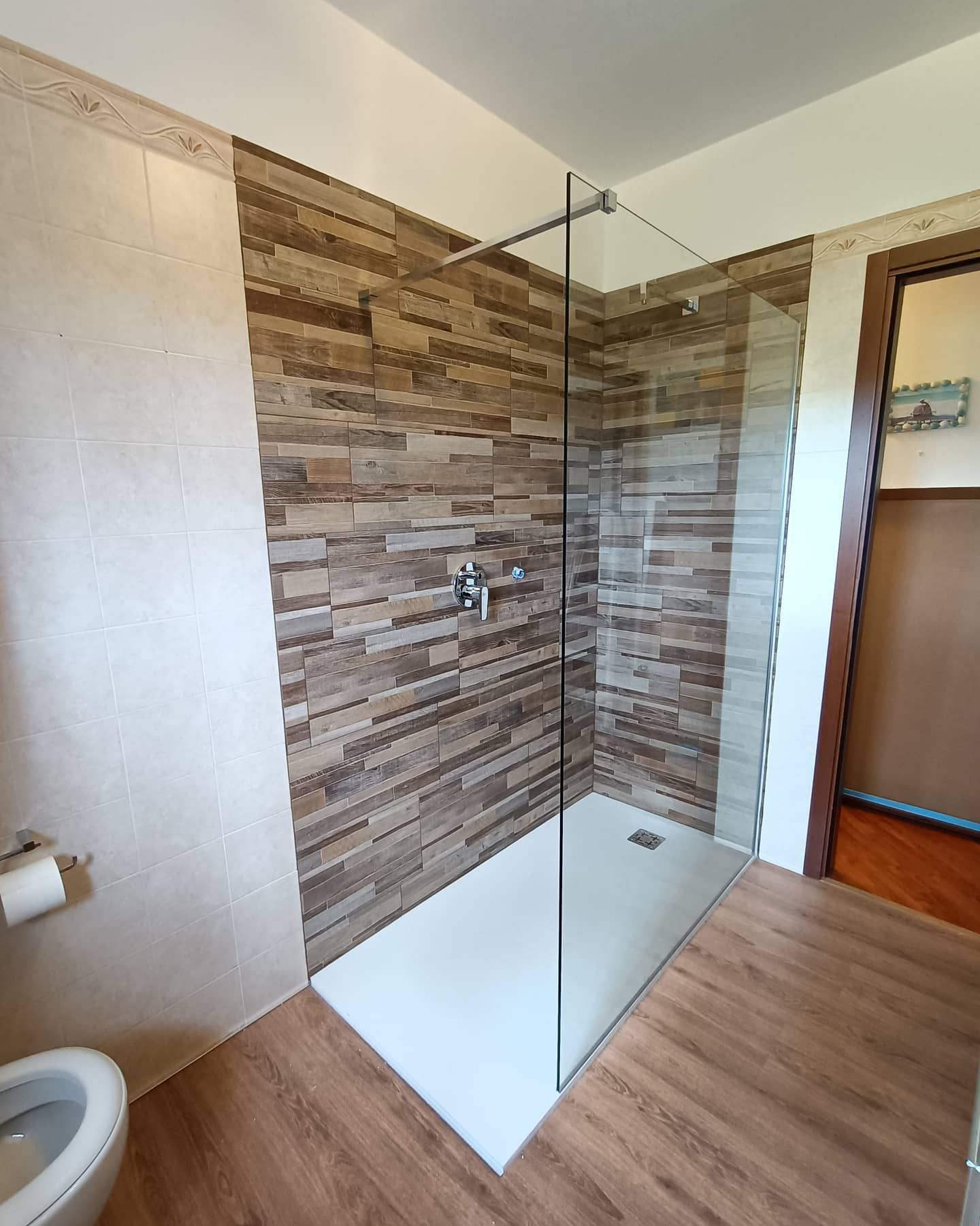 Bagno moderno con box doccia in vetro, parete piastrellata effetto legno, piatto doccia bianco e WC.
