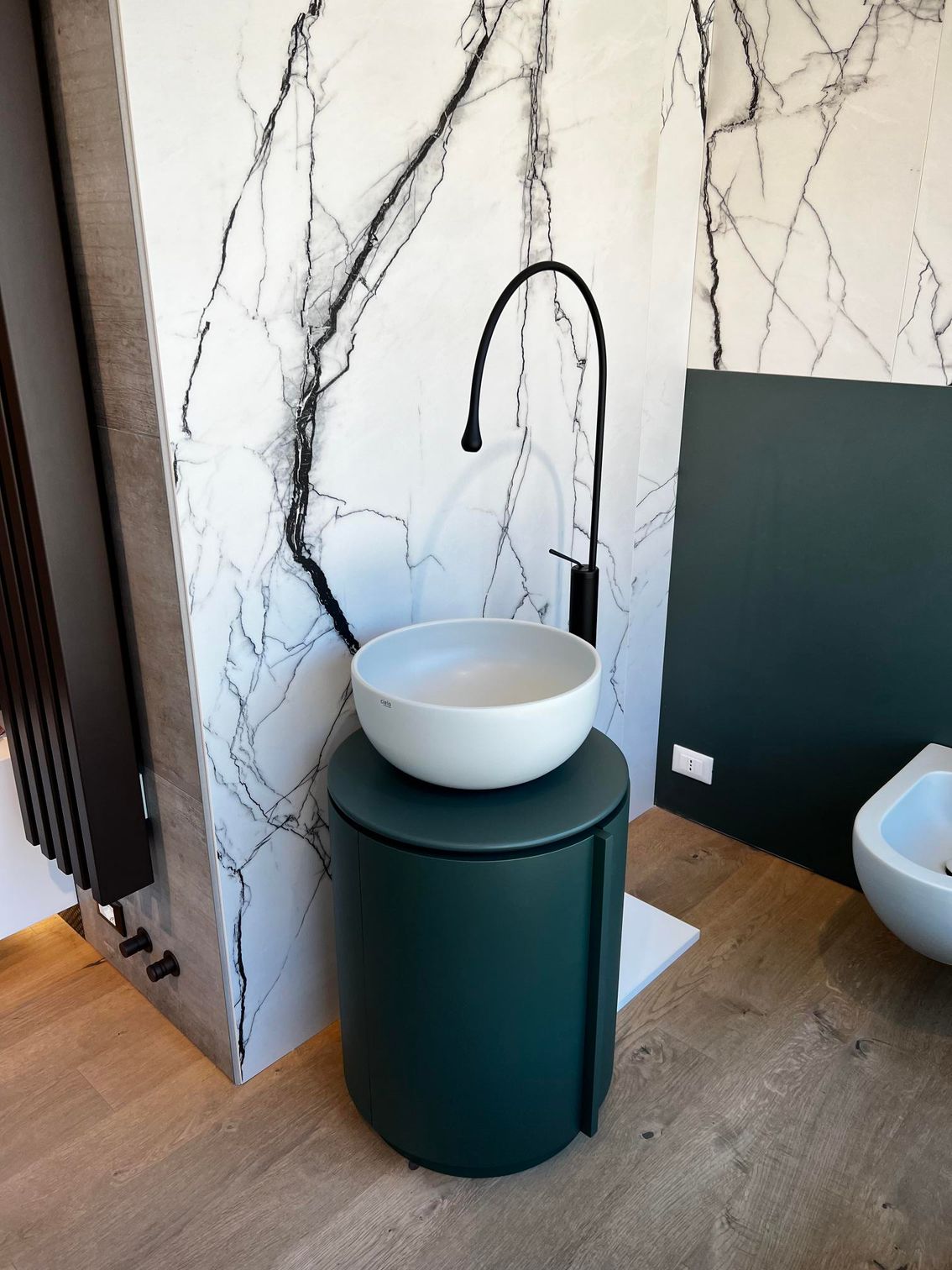 Bagno moderno con lavandino rotondo bianco su piedistallo verde scuro, contro una parete con motivi in ​​marmo.