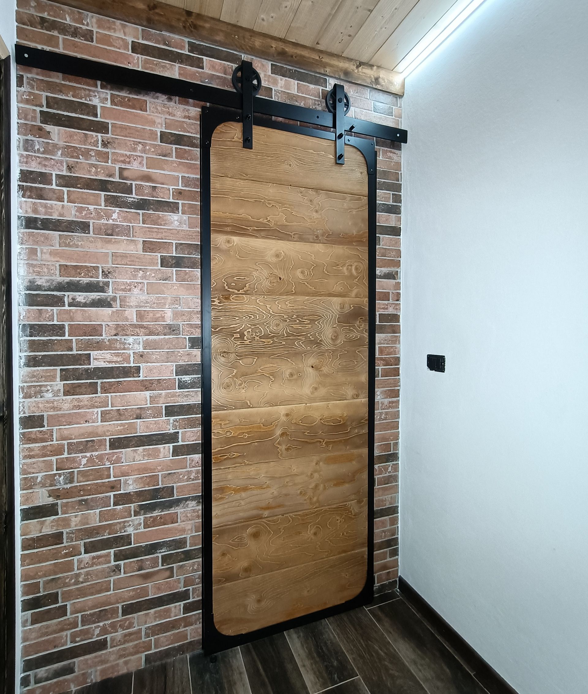 Porta del fienile su muro di mattoni, telaio nero, porta in legno, ferramenta nera, finiture in legno, muro bianco.