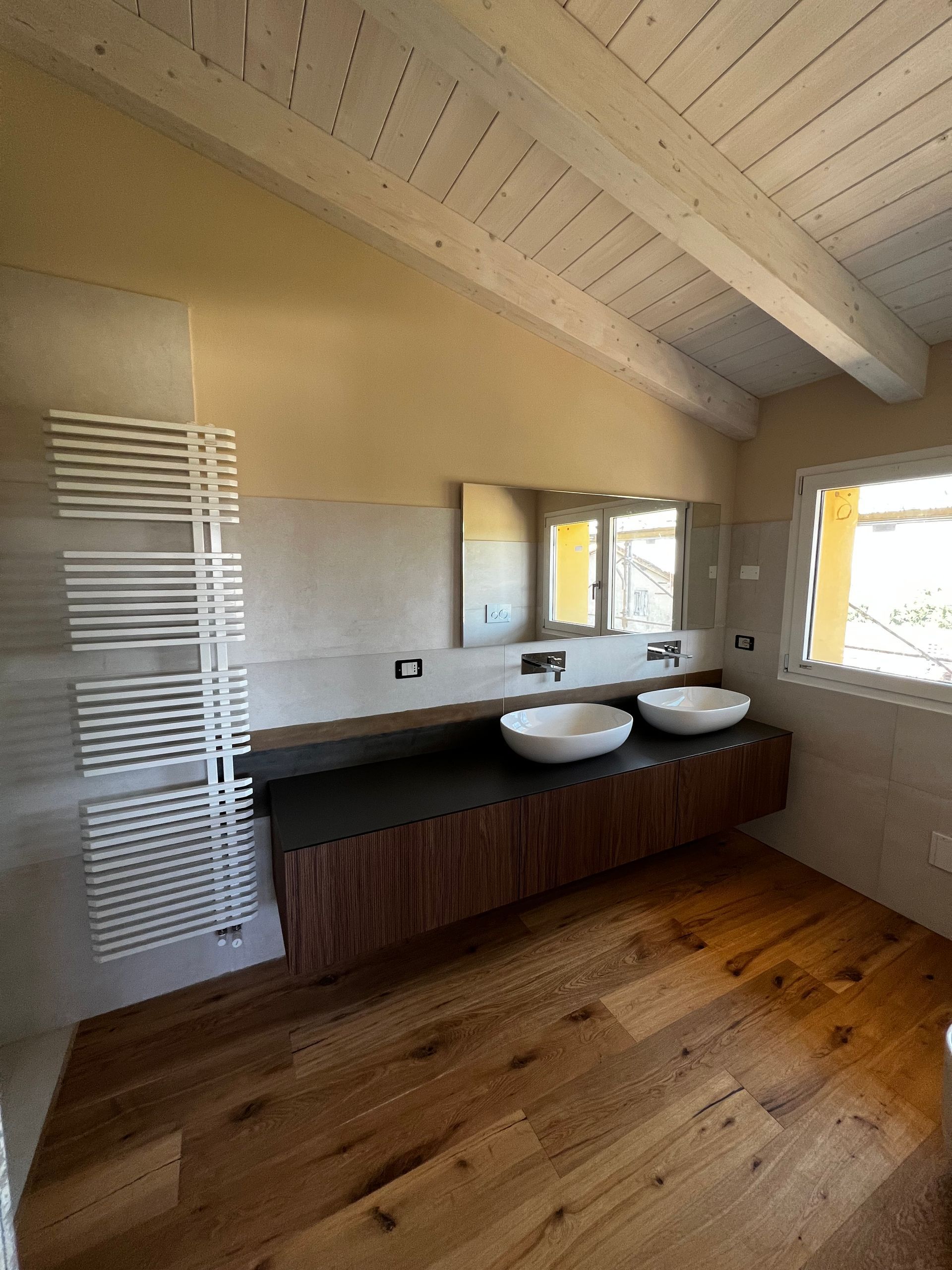 Bagno con pavimento in legno, lavandini bianchi e un lungo mobiletto in legno scuro. Portasciugamani bianco.