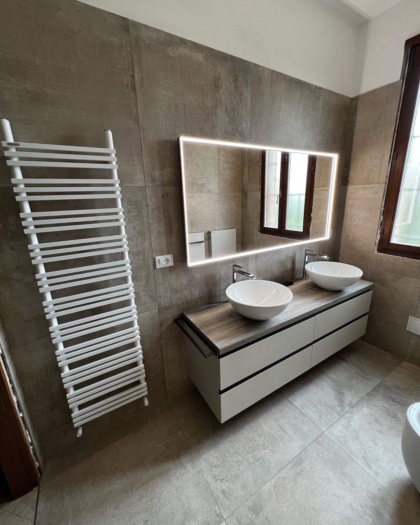 Bagno moderno con lavandini bianchi su un mobile, uno specchio illuminato e uno scaldasalviette.