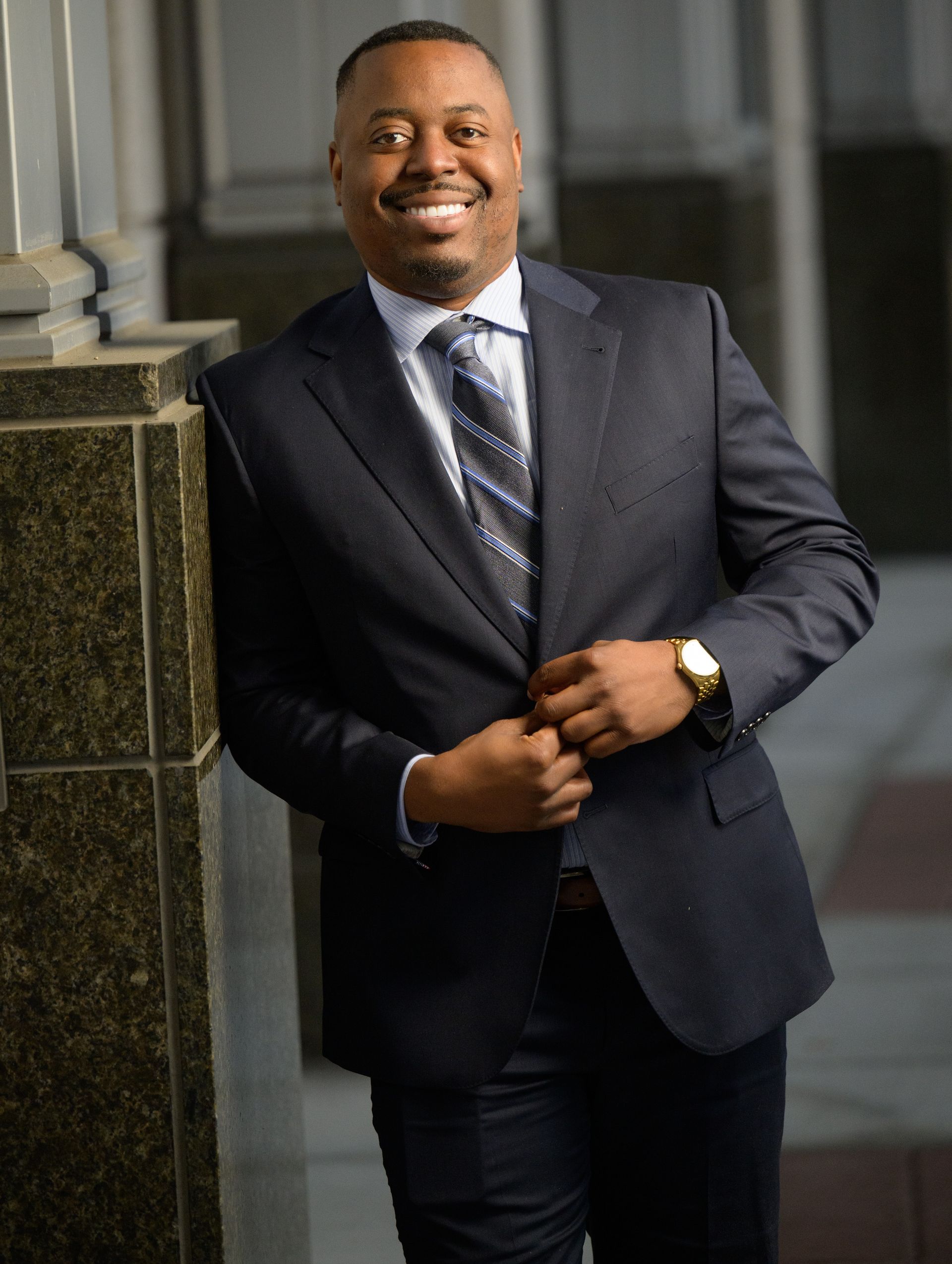 David Alcius in a dark suit and patterned tie standing outdoors, leaning against a stone pillar in Orlando, Florida