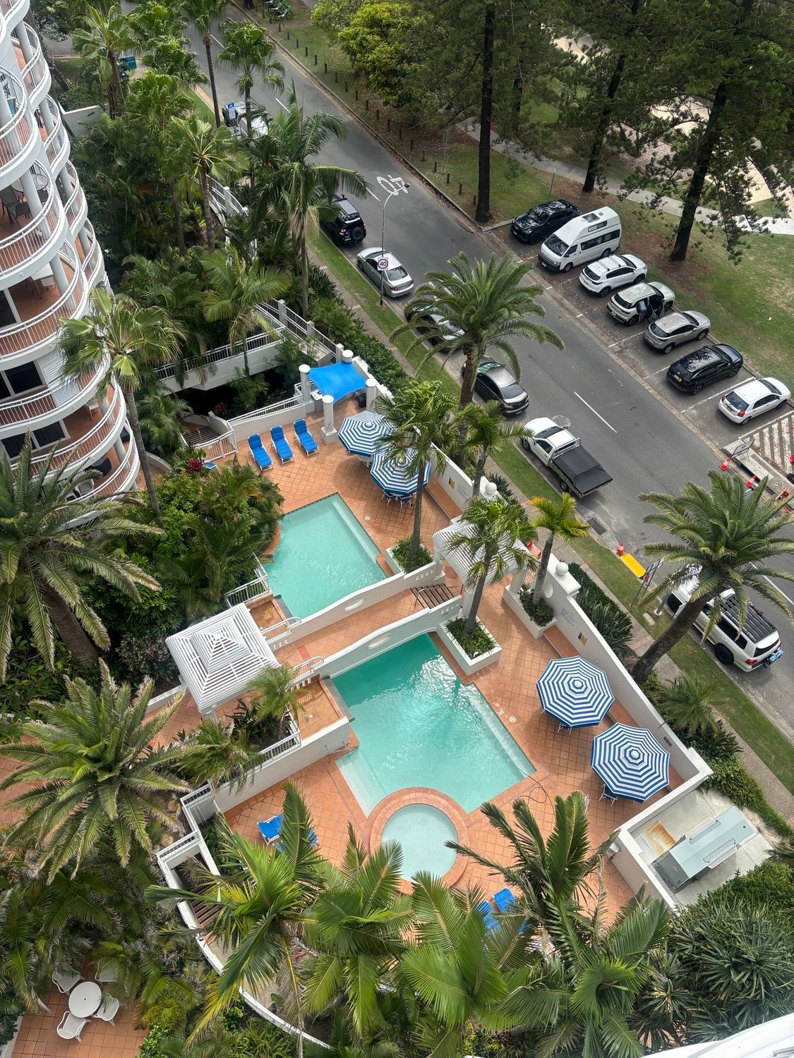 Apartment pool courtyard with turquoise pools, palm trees, and parked cars along the street