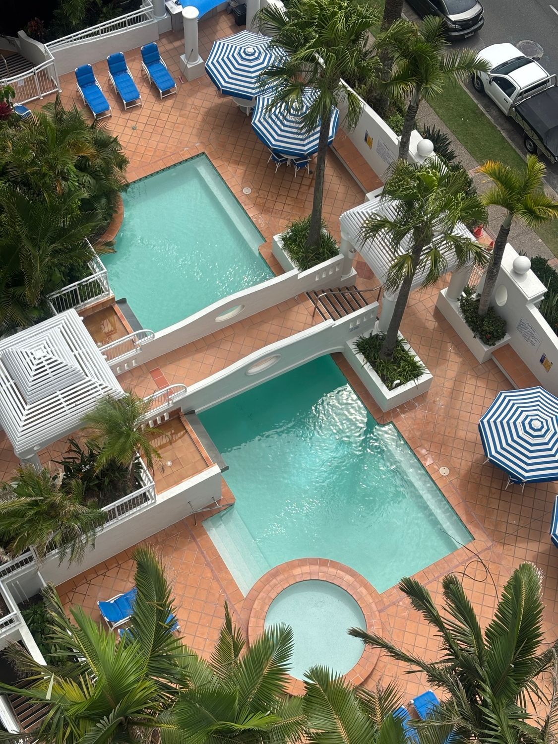 Overhead view of two turquoise hotel pools with tan deck, blue loungers, umbrellas, and palm trees