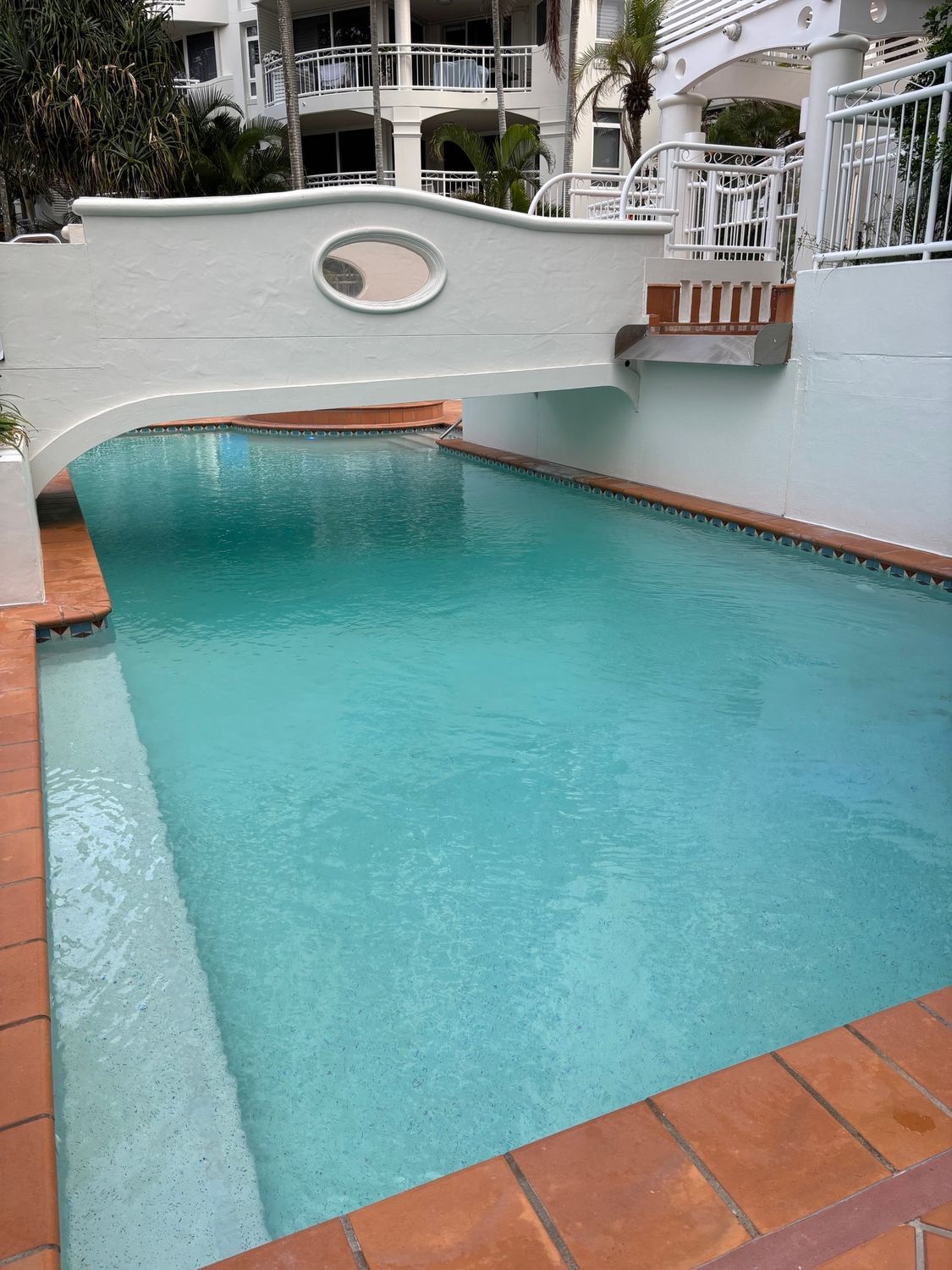Turquoise swimming pool with terracotta tile edge and white stucco wall, viewed from a patio.