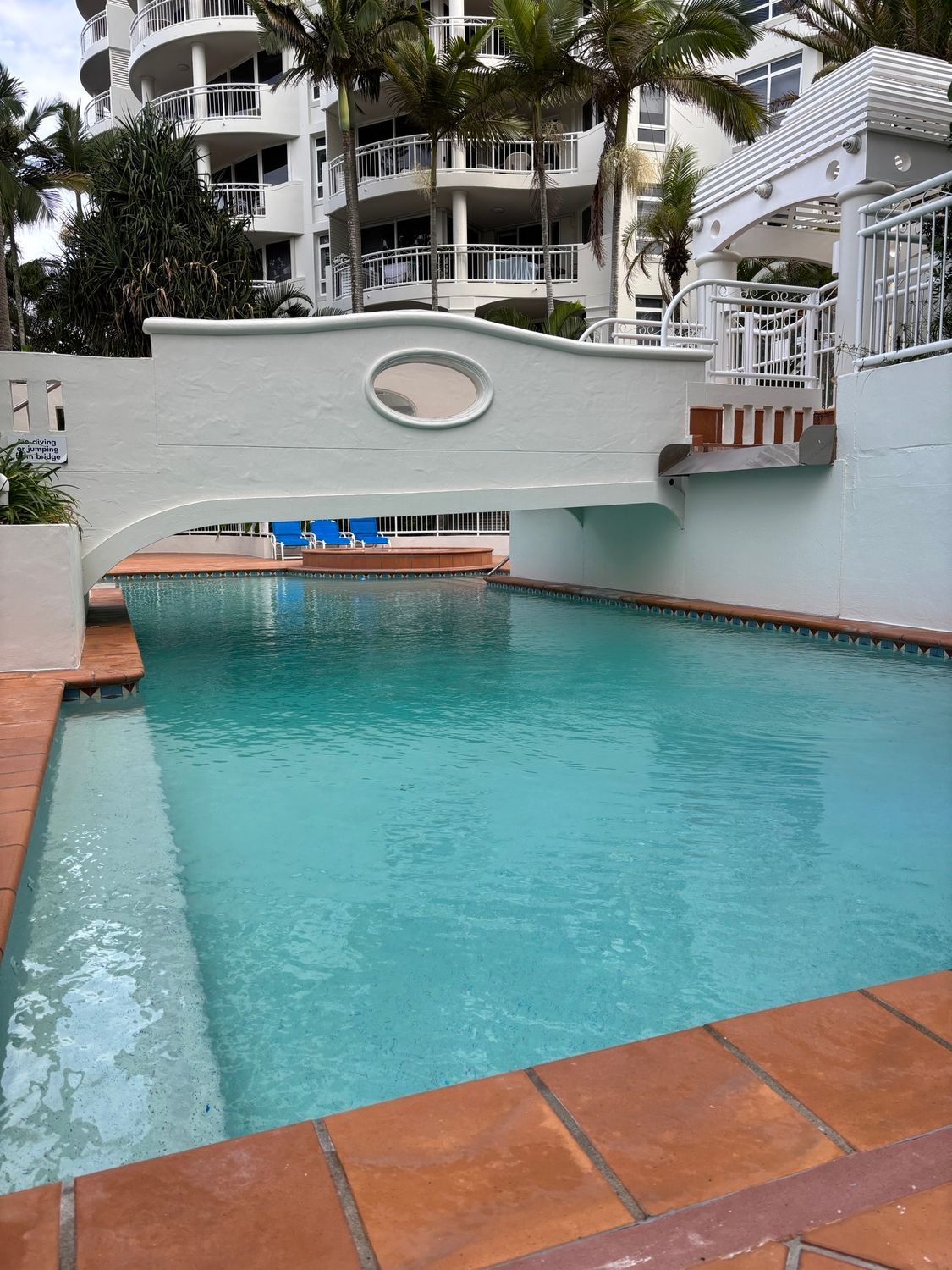 Outdoor turquoise swimming pool with a white bridge and apartment buildings in the background