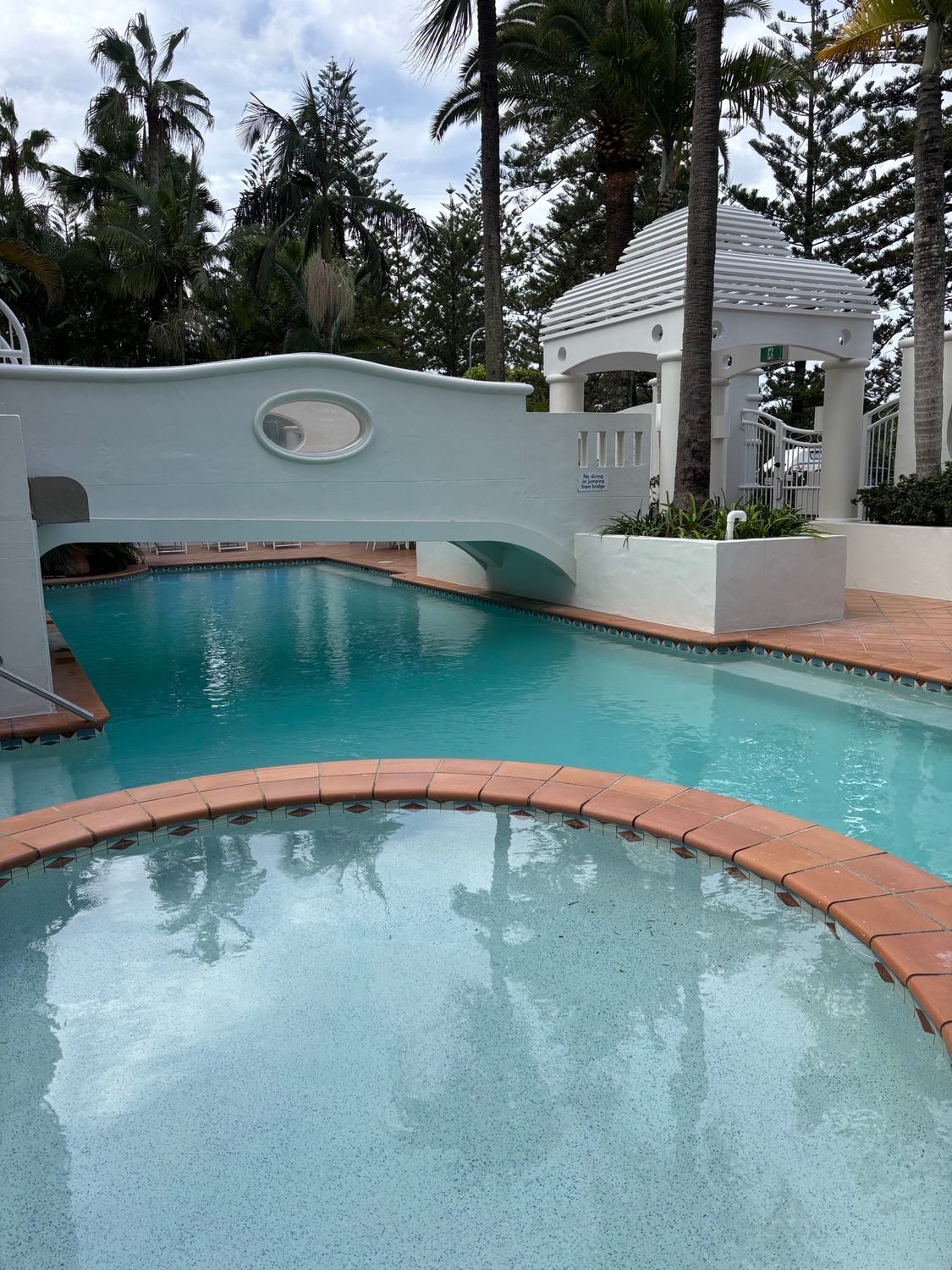 Resort pool with turquoise water, a curved bridge, and a white gazebo surrounded by tropical trees