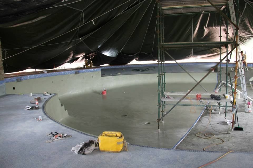 Indoor construction site with a large curved concrete pool or basin under a tarp-covered roof.