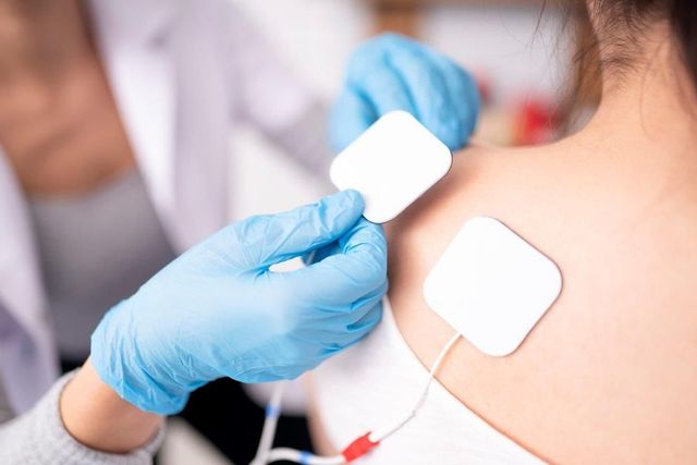 Persona con guanti blu che applica elettrodi alla spalla di una donna per la terapia fisica.