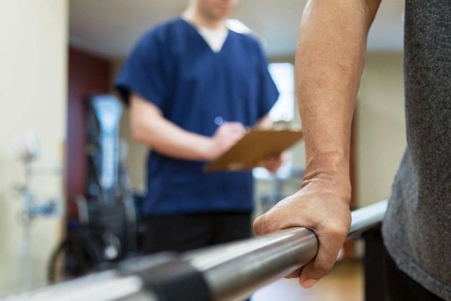 Persona che utilizza un corrimano per sostenersi durante la terapia fisica, con un medico sullo sfondo che prende appunti.