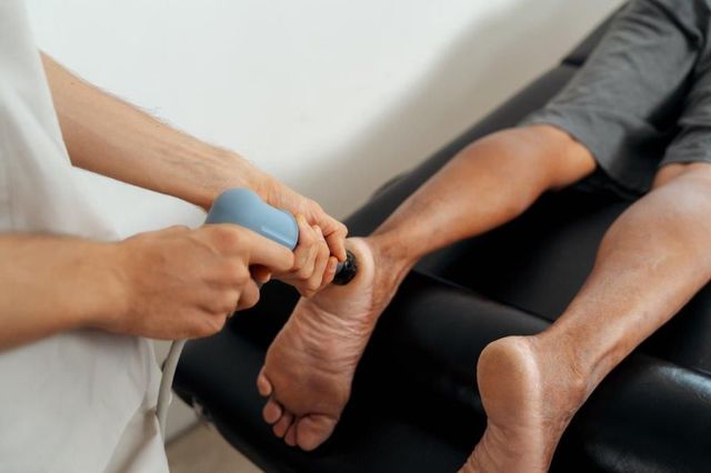 Una persona riceve una terapia plantare con uno strumento massaggiante blu. Parete bianca e sedia nera.