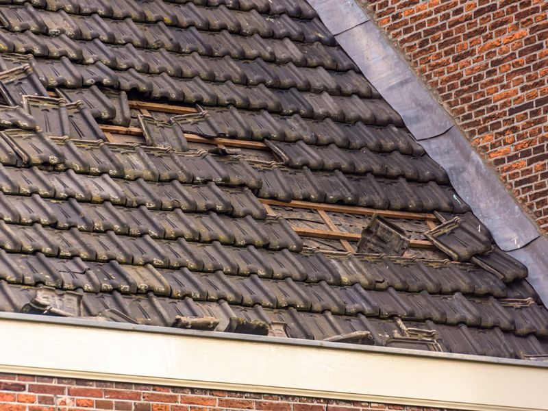 Damaged tiled roof with missing and broken shingles near a brick chimney.