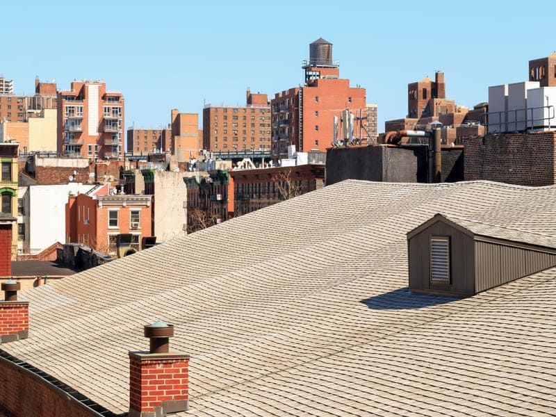 City rooftops under a clear blue sky, with brick buildings and a large gray rooftop in the foreground