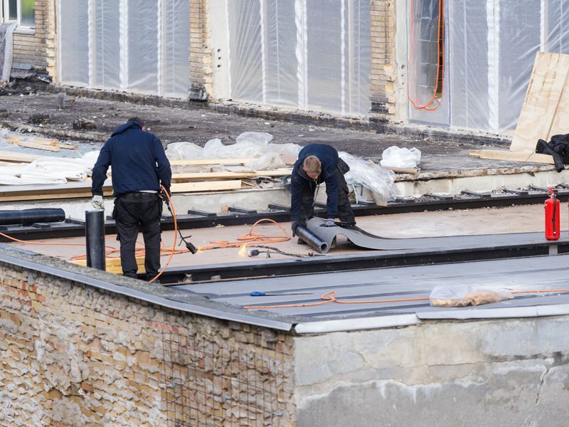 Two workers repairing a rooftop near a brick building, with tools and a red fire extinguisher nearby.