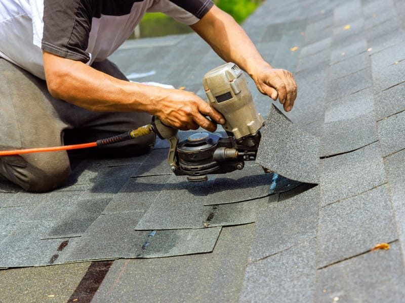 Worker using a nail gun to install gray asphalt shingles on a roof