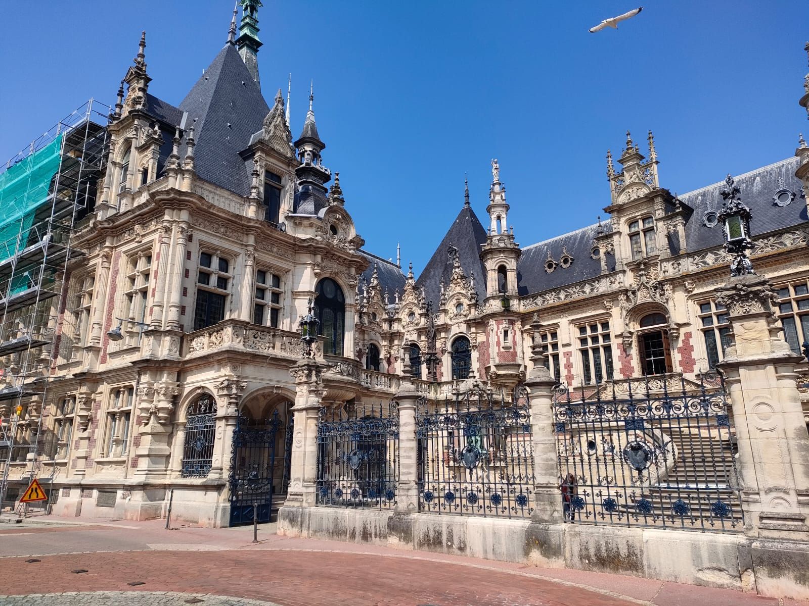 Bâtiment en pierre orné, avec un toit sombre et une grille en fer forgé, se détachant sur un ciel d'un bleu éclatant.
