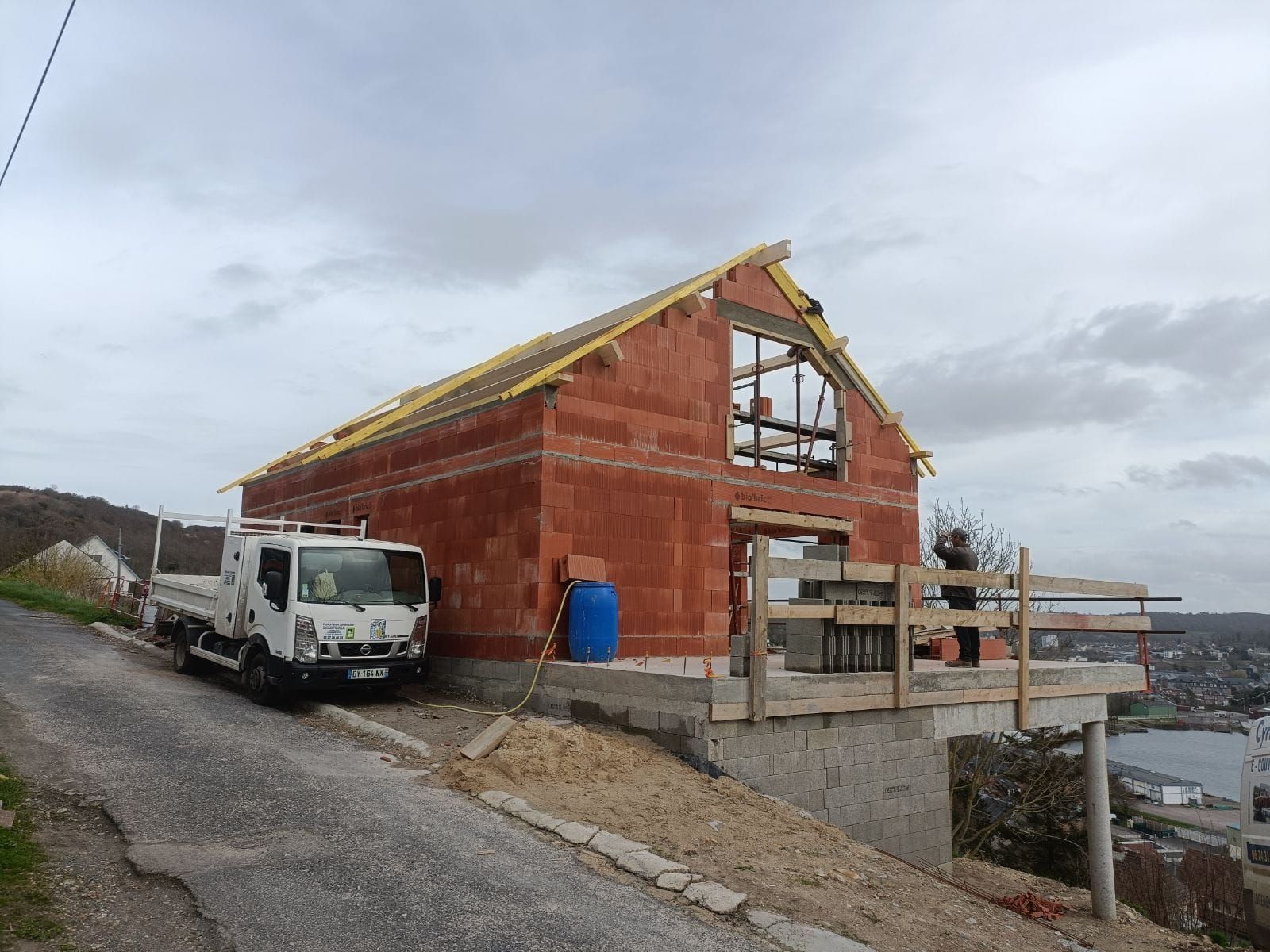 Bâtiment en construction : structure en briques rouges à flanc de colline surplombant l'eau, avec un camion garé à côté.