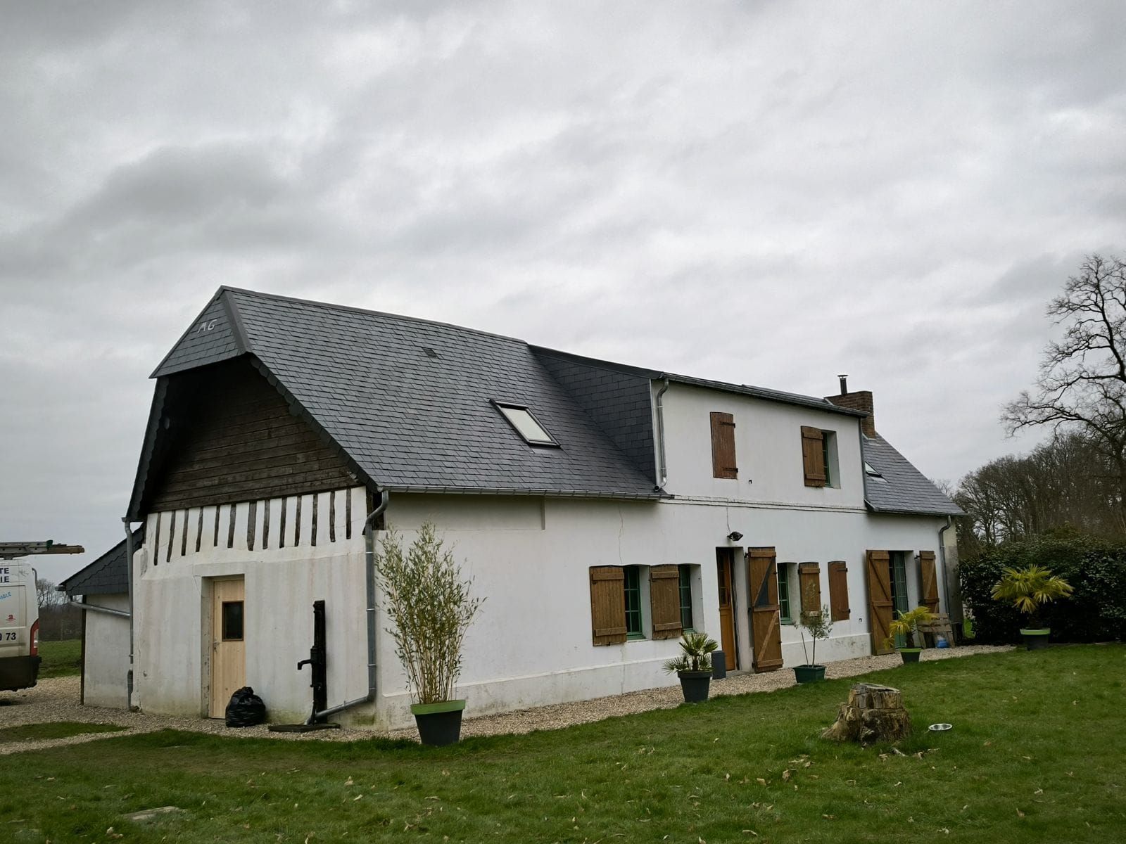 Maison blanche au toit sombre et aux volets marrons sous un ciel nuageux.