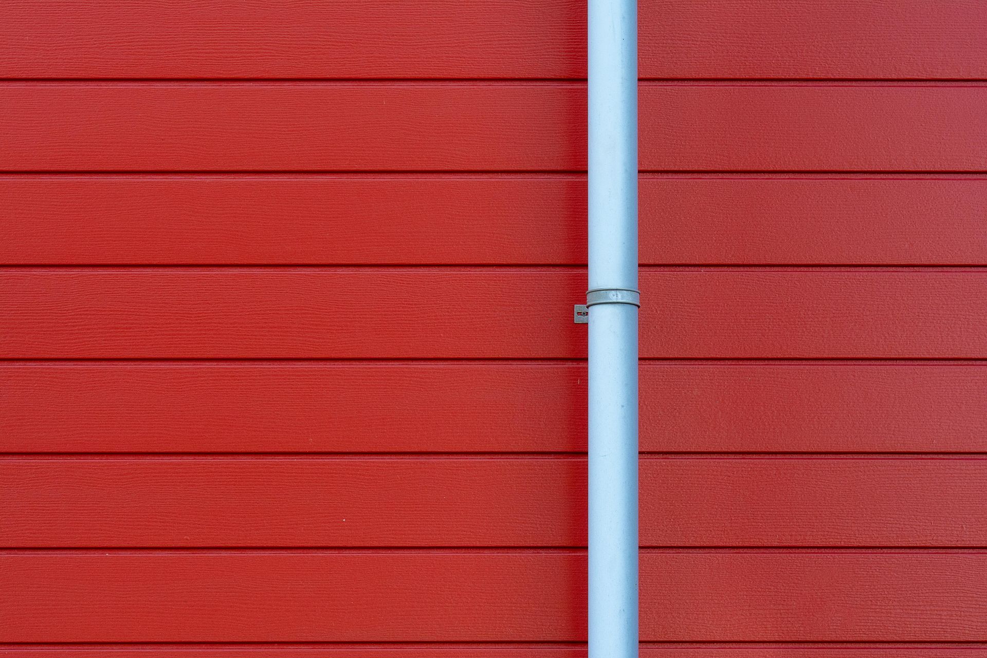 Tuyau argenté fixé à un mur rouge recouvert de planches horizontales.