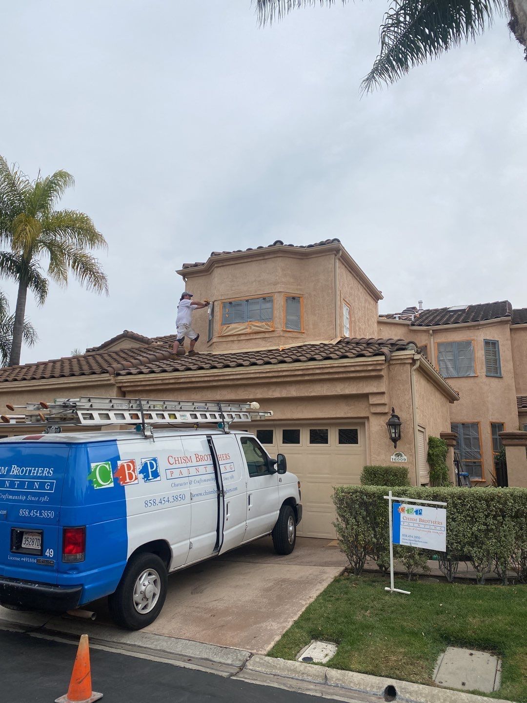 A white van parked near a house with a person on the roof. The van has a ladder.