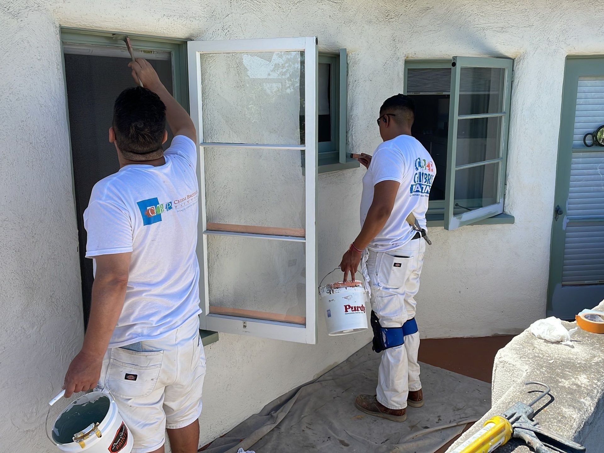 Two painters in white clothing painting window frames on a stucco building.