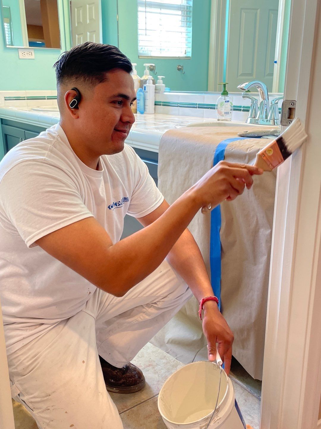 A person painting a door frame in a bathroom, wearing white overalls, holding a paintbrush and paint bucket.