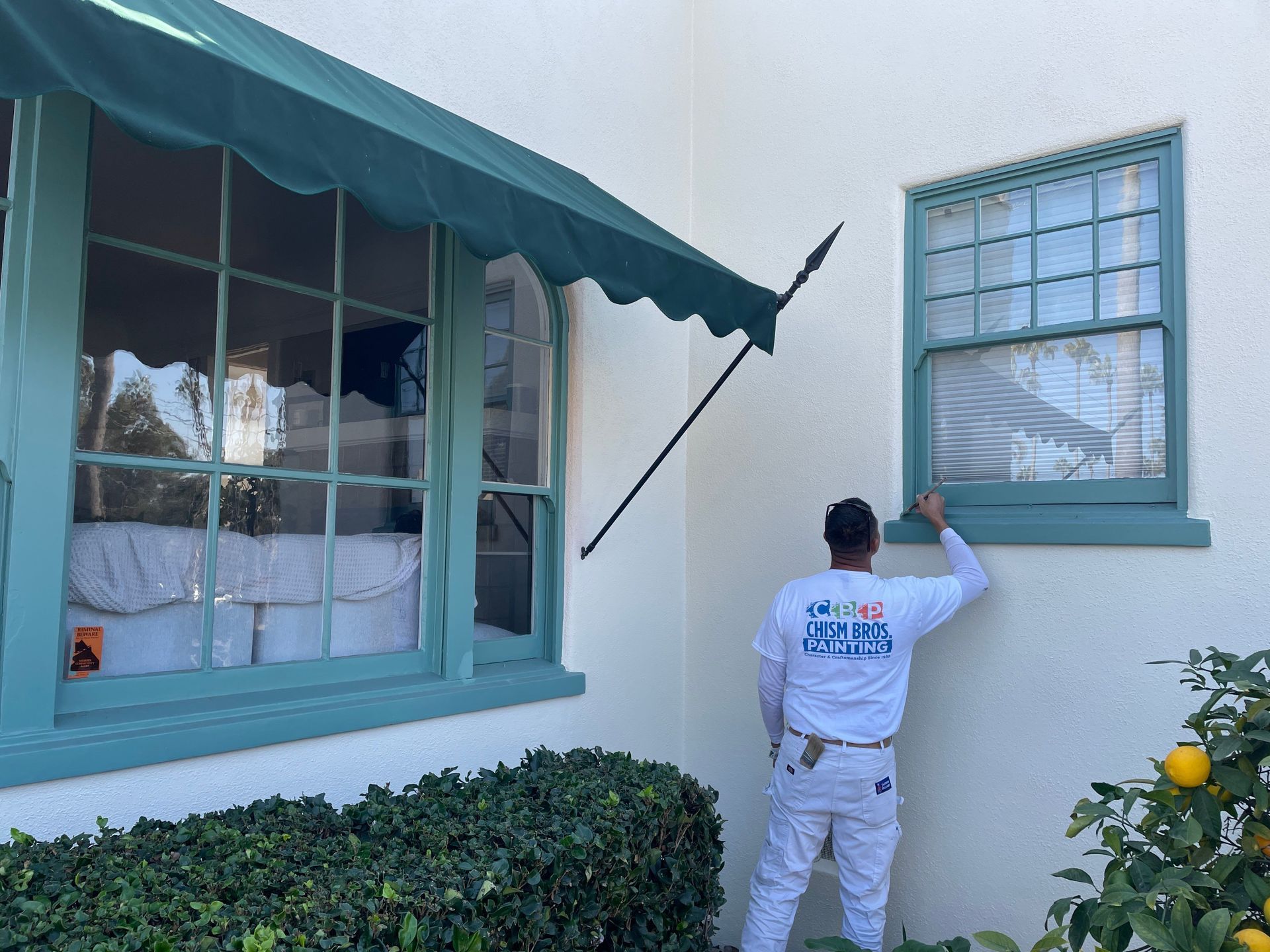 Person painting window trim, teal on white building. Green awning and foliage.