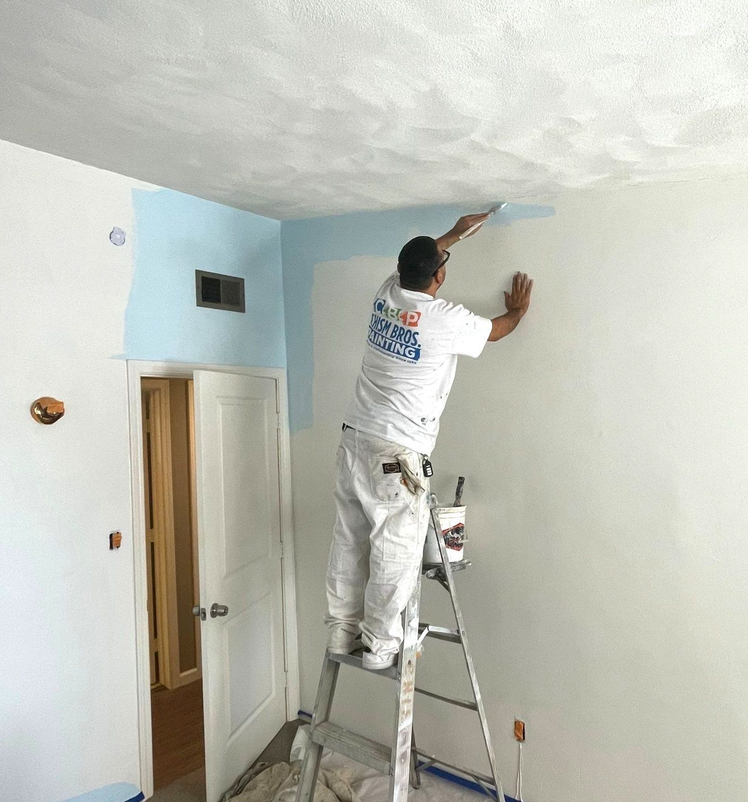 Painter on a ladder applies blue paint to a wall's edge, working in a room.