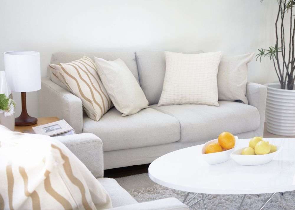 A living room with a white sofa and pillows, a round coffee table with fruit, and a lamp.