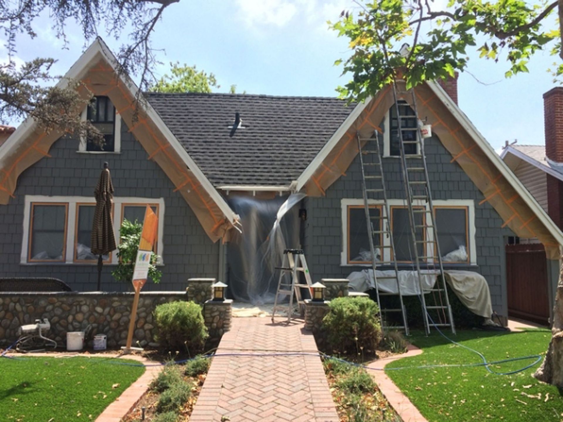 House exterior being painted, blue-gray siding, brown roof, ladders, plastic sheeting, brick path, landscaping.
