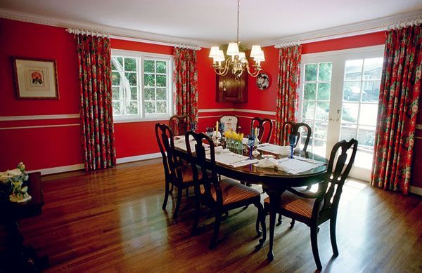 Red dining room with oval table, chairs, chandelier, and floral curtains.