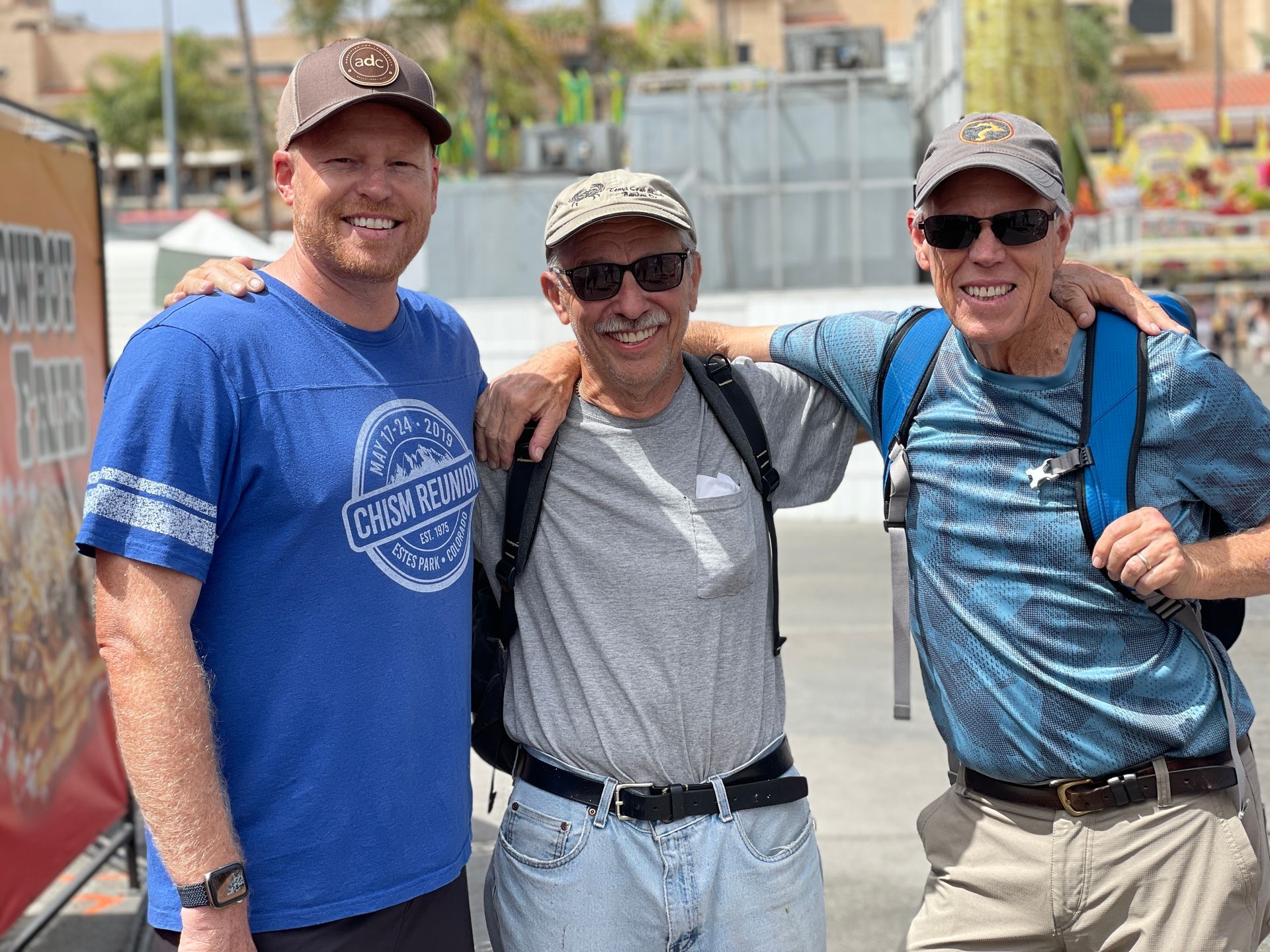 Three men posing outdoors, two with arms around the center man. All wearing hats, smiling.