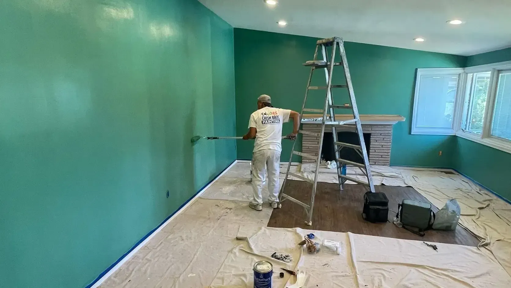 A person paints a room teal. Paint cans and drop cloths are on the floor.