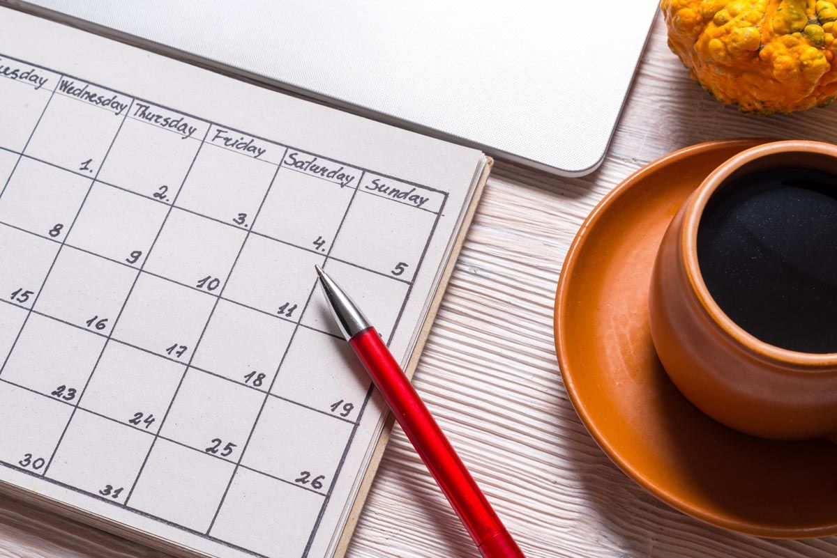 Calendar and red pen on notebook with coffee and laptop on table.