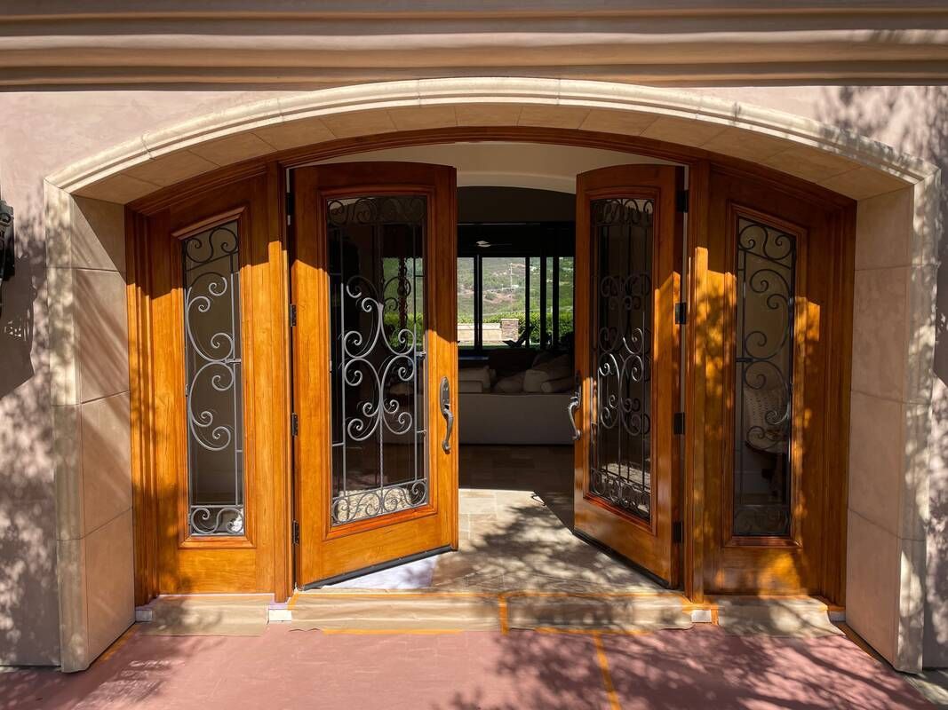 Wooden double doors with glass and ornate ironwork, arched entryway, open to a bright interior.