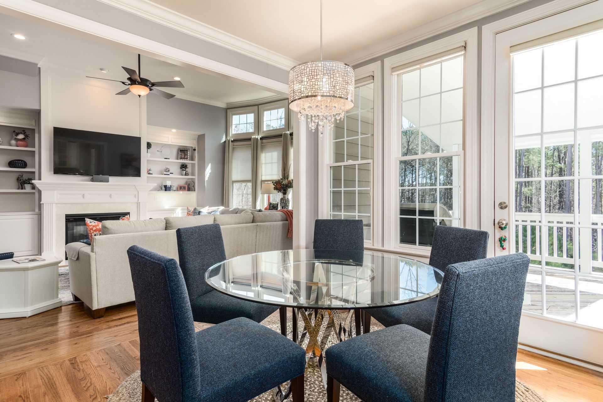 Dining room with a glass table, blue chairs, and a chandelier, with a view of a living area and large windows.
