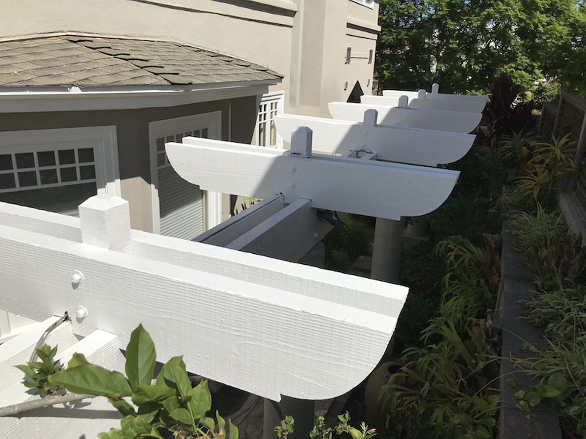 White pergola attached to a light-colored building, with plants along the side.