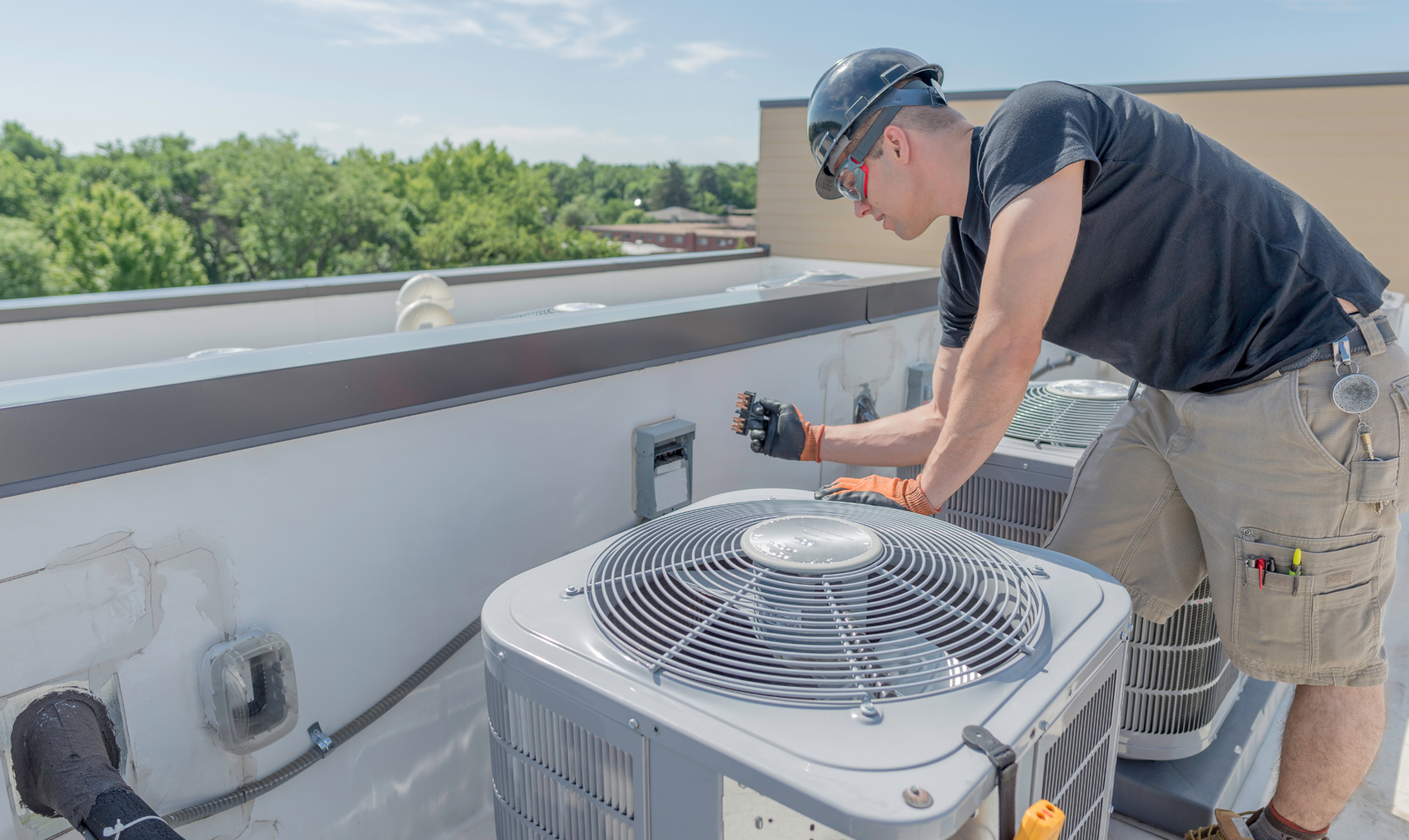 Technician on a rooftop adjusting a rooftop HVAC unit beside a white parapet wall.