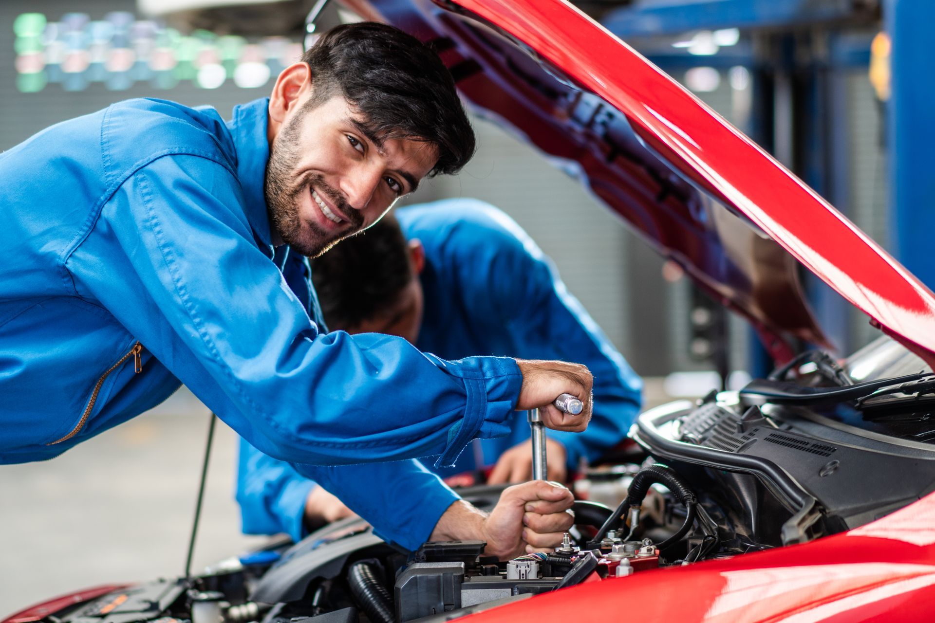 two mechanics work on a red car with the hood open