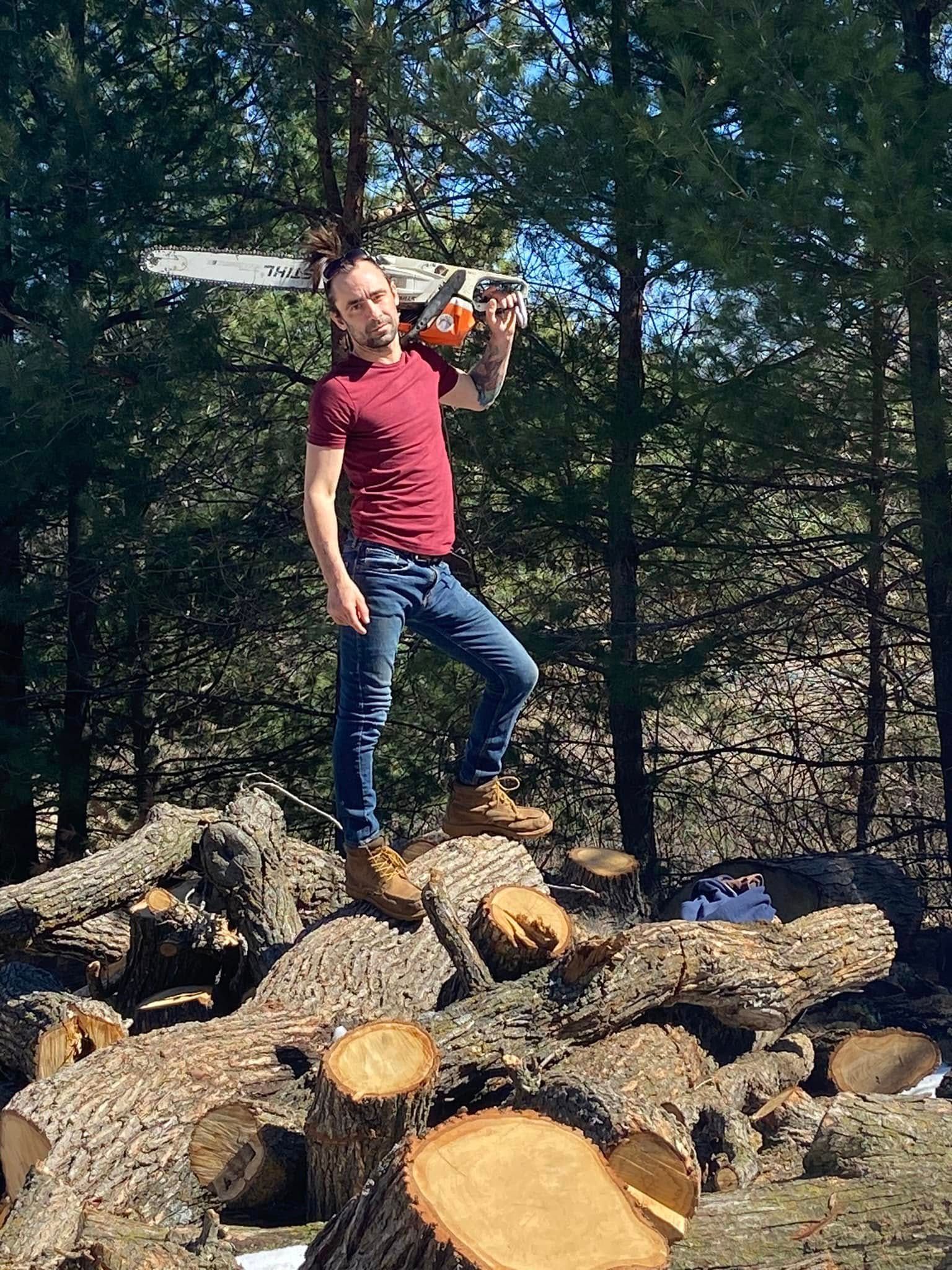 Person drinking from a bottle while standing on stacked cut logs outdoors in a wooded area