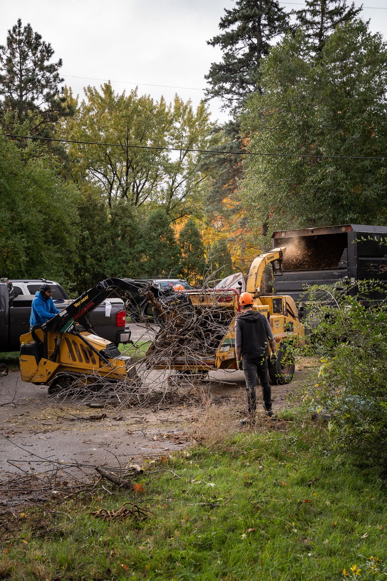 Two workers clear debris with a yellow skid steer in a muddy yard near trees and parked trucks.
