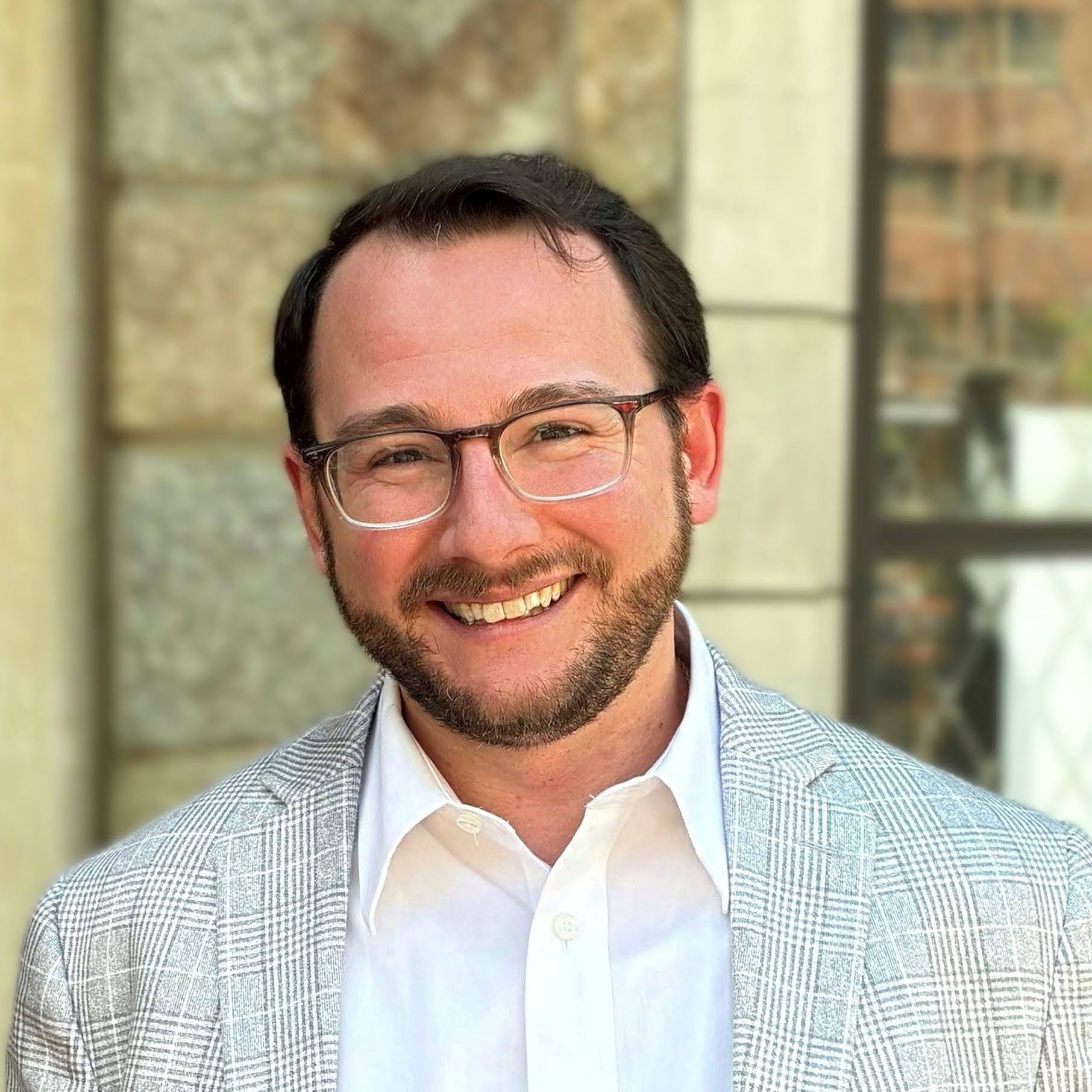 Man with glasses and beard, wearing a suit, smiles in front of a dark background.