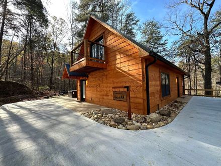 Wooden cabin with balcony on a concrete driveway, surrounded by bare trees in a wooded area.