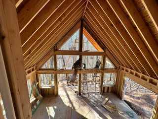 A wooden cabin loft with A-frame beams, large windows, and sunlight on the floor and walls.