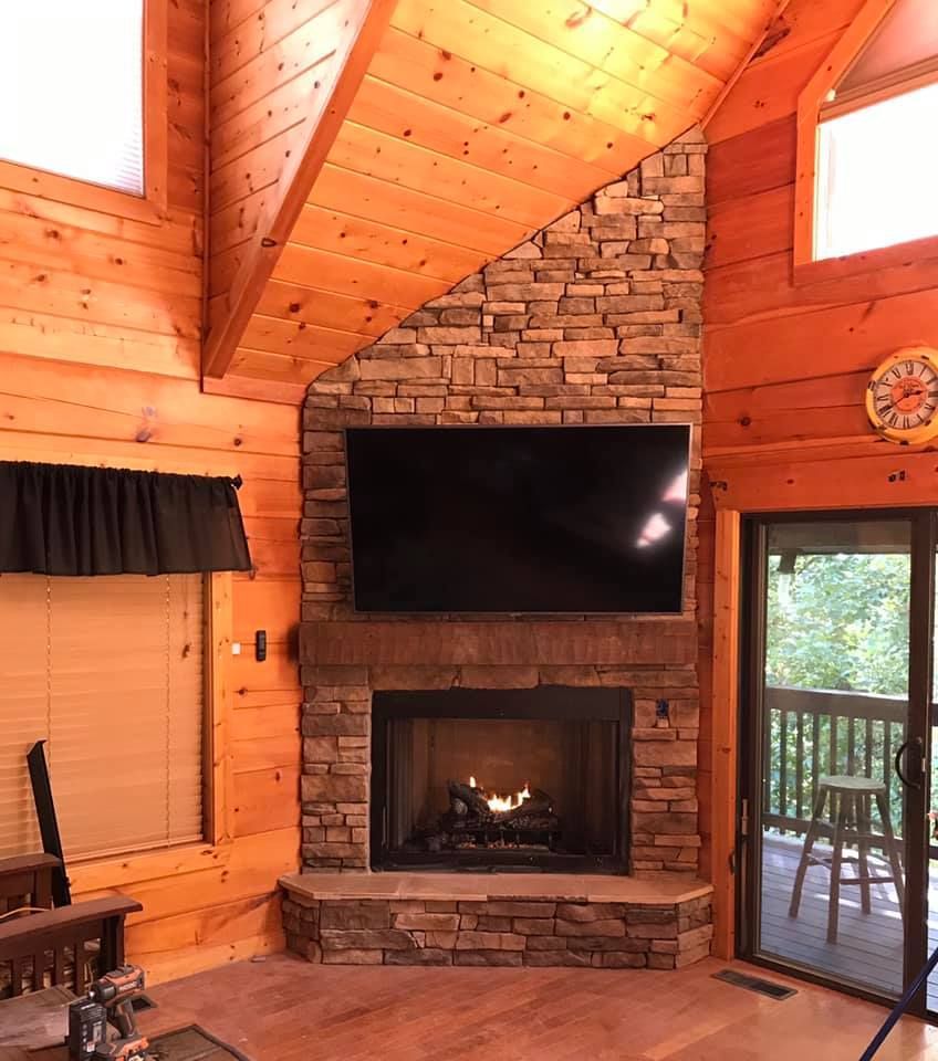 Cozy cabin living room with stone fireplace, mounted TV, and wood-paneled walls with a vaulted ceiling